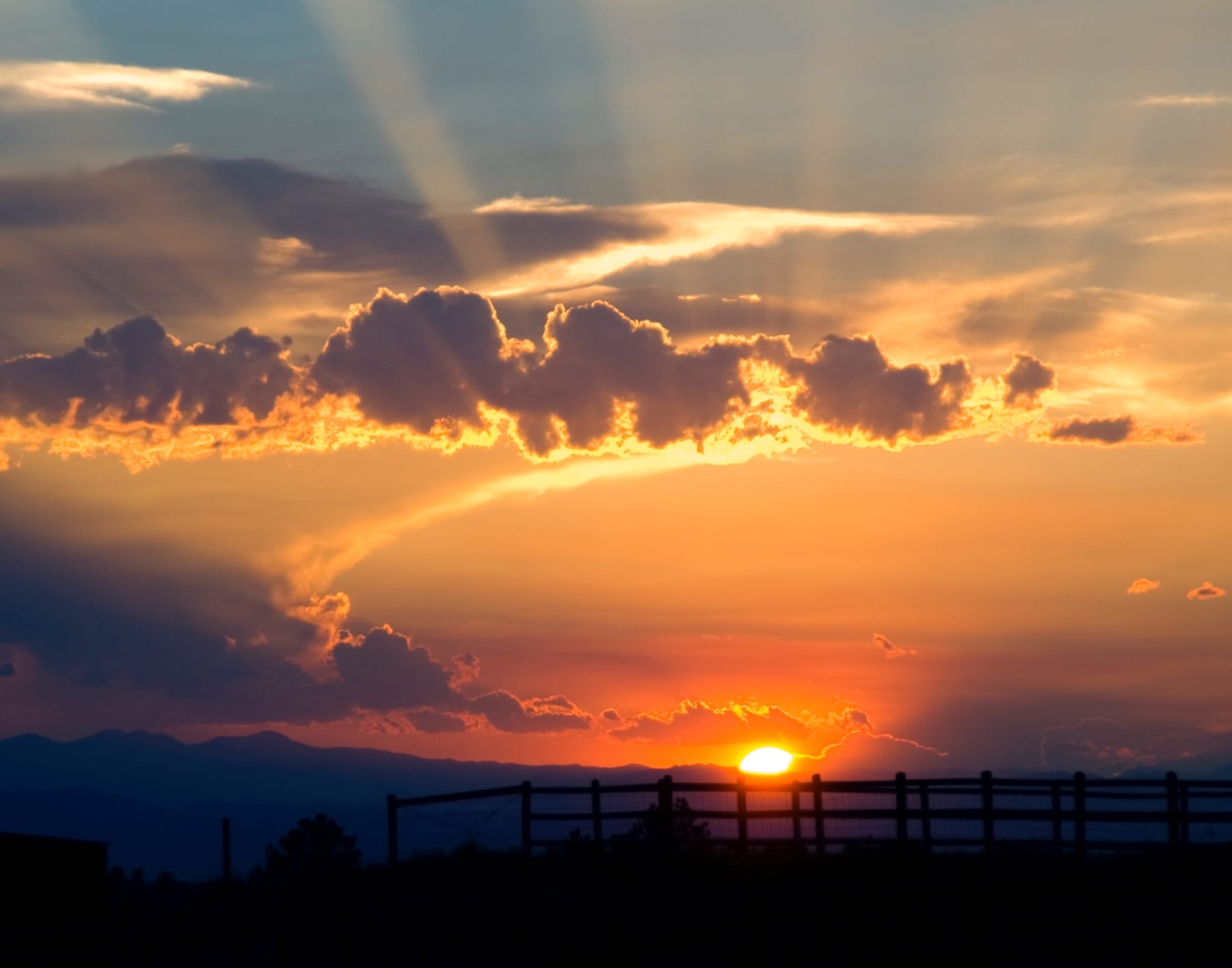 The sun setting behind the pasture fence, with the Rocky Mountains on the horizon